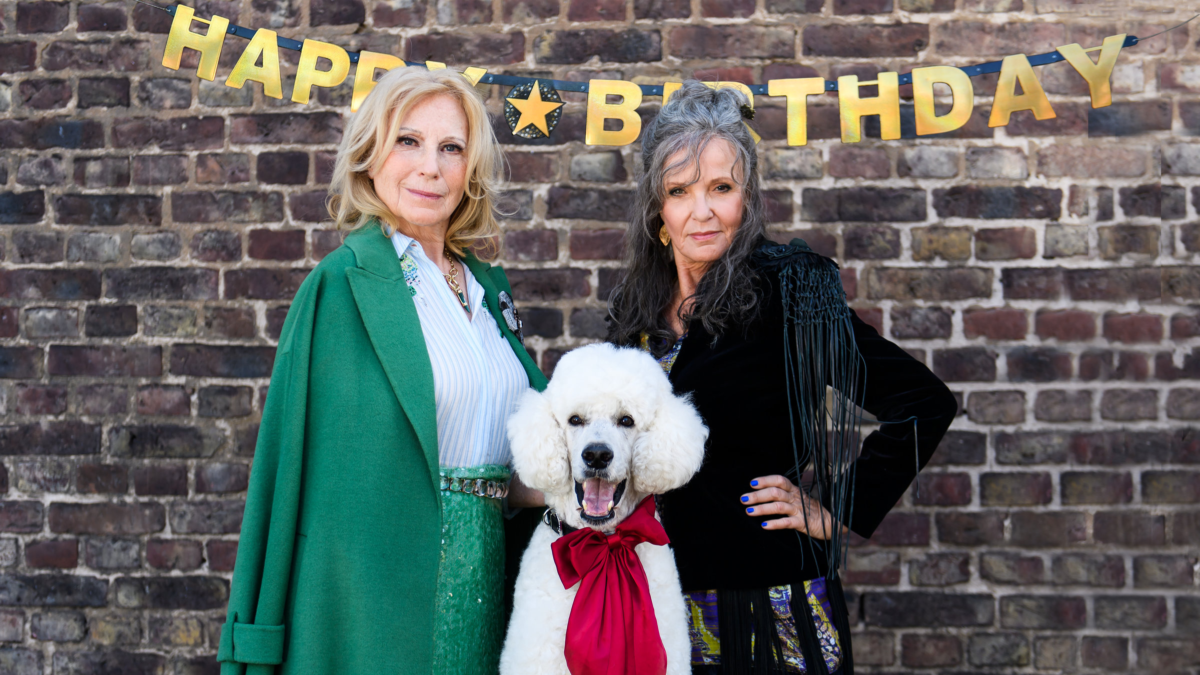"Mona und Marie - Ein etwas anderer Geburtstag": Mona (Maren Kroymann) steht perfekt zurechtgemacht in einem offenen grünen Mantel über einer weißen Bluse vor einer Backsteinwand. Ihre Schwester Marie (Ulrike Kriener) schaut selbstbewusst in einer braunen Fransenlederjacke in die Kamera. Zwischen ihnen ein sitzt  ein weißer Königspudel mit offenem Maul und einer roten dicken Schleife um den Hals. Über dem Bild an der Backsteinmauer hängt eine goldene Druckschriftgirlande "Happy Birthday".