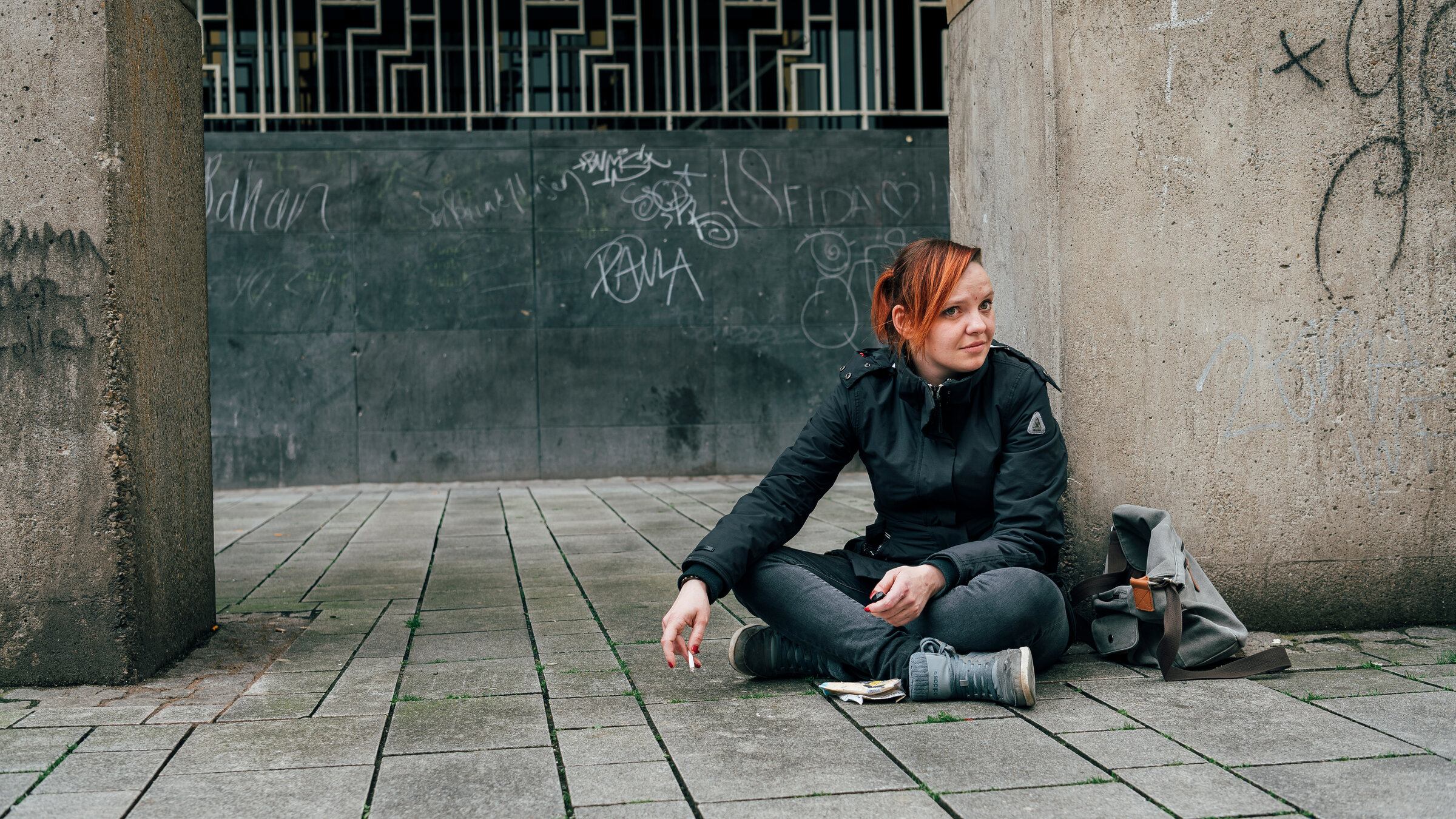 "37°: Schutzlos ausgeliefert - Frauen zwischen Straße und Notunterkunft": Leonie sitzt im Schneidersitz auf dem Boden vor einem Betonpfeiler. Sie hat rote Haare und trägt schwarze Kleidung. Neben ihr liegt eine Tasche auf dem Boden.