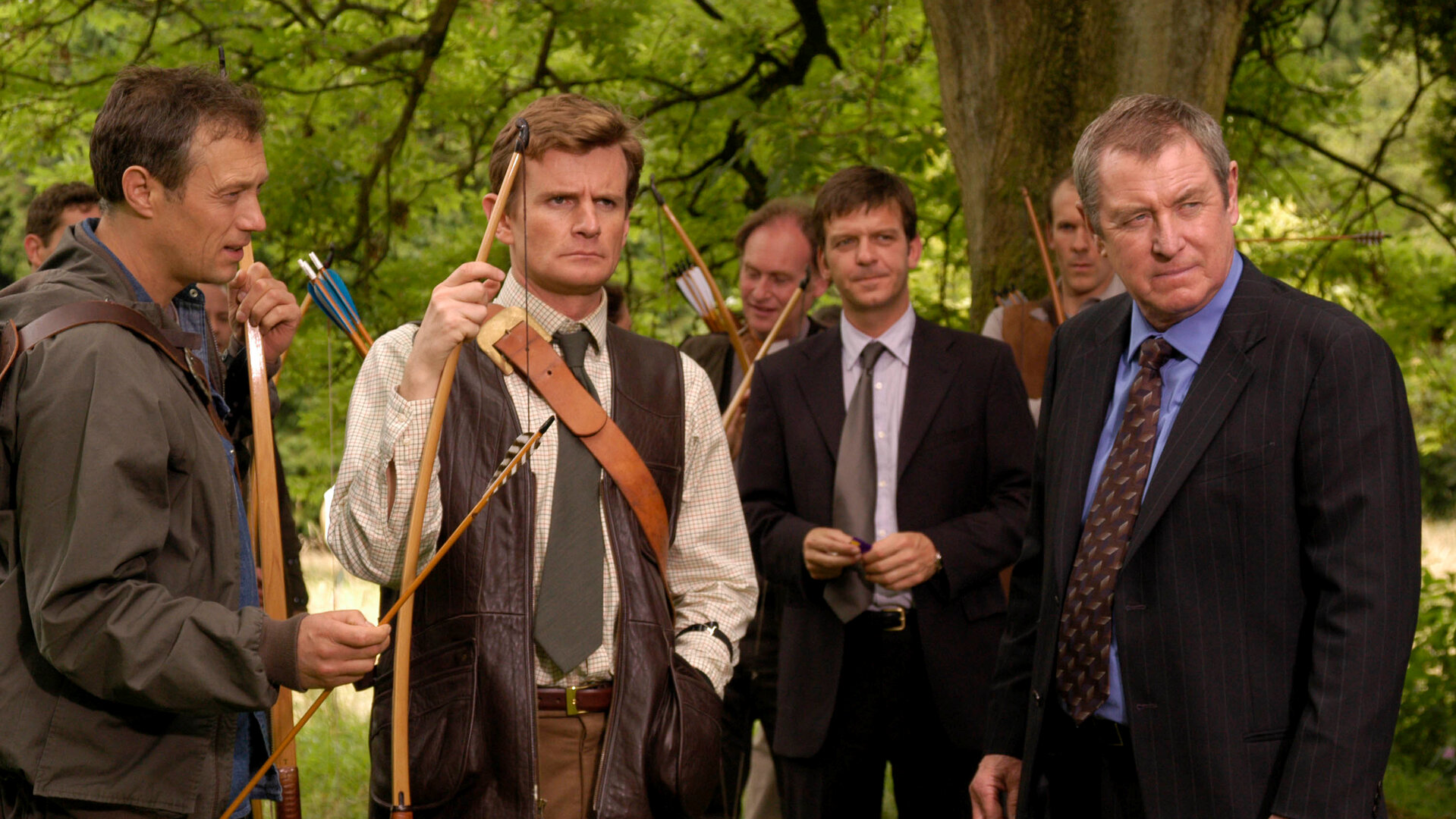 "Inspector Barnaby - Ganz in Rot": Robin Lawson (Martin Wenner), und Ned Fitzroy (Charles Edwards) mit Pfeil und Bogen in den Händen stehen zusammenmit DS Ben Jones (Jason Hughes) und DCI Tom Barnaby (John Nettles) unter einem Baum und schauen in eine Richtung. Hinter ihnen stehen noch weitere Männer.