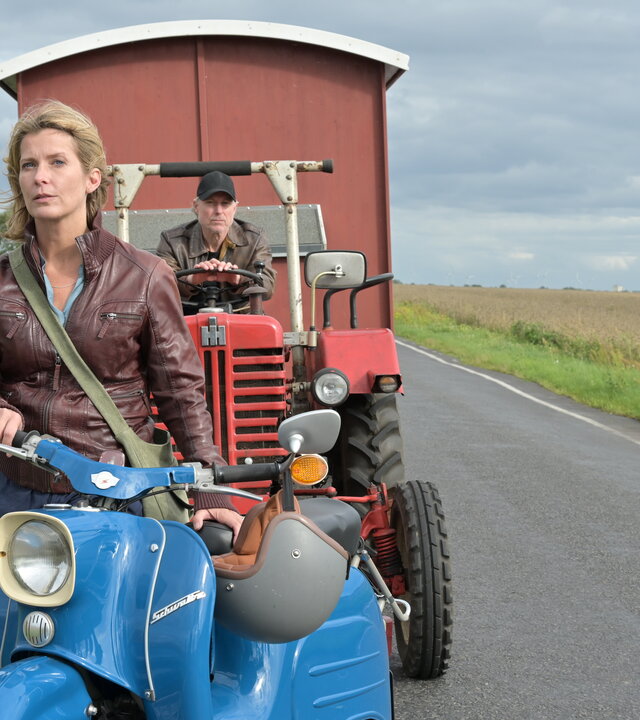 "Nächste Ausfahrt Glück - Übers Ziel hinaus": Katharina (Valerie Niehaus) steht mit dem Helm in der Hand neben ihrem Moped, einer "Schwalbe", auf einer Landstraße. Hinter ihr sitzt Juri (Dirk Borchardt) auf einem Traktor, der einen Schäferwagen zieht.