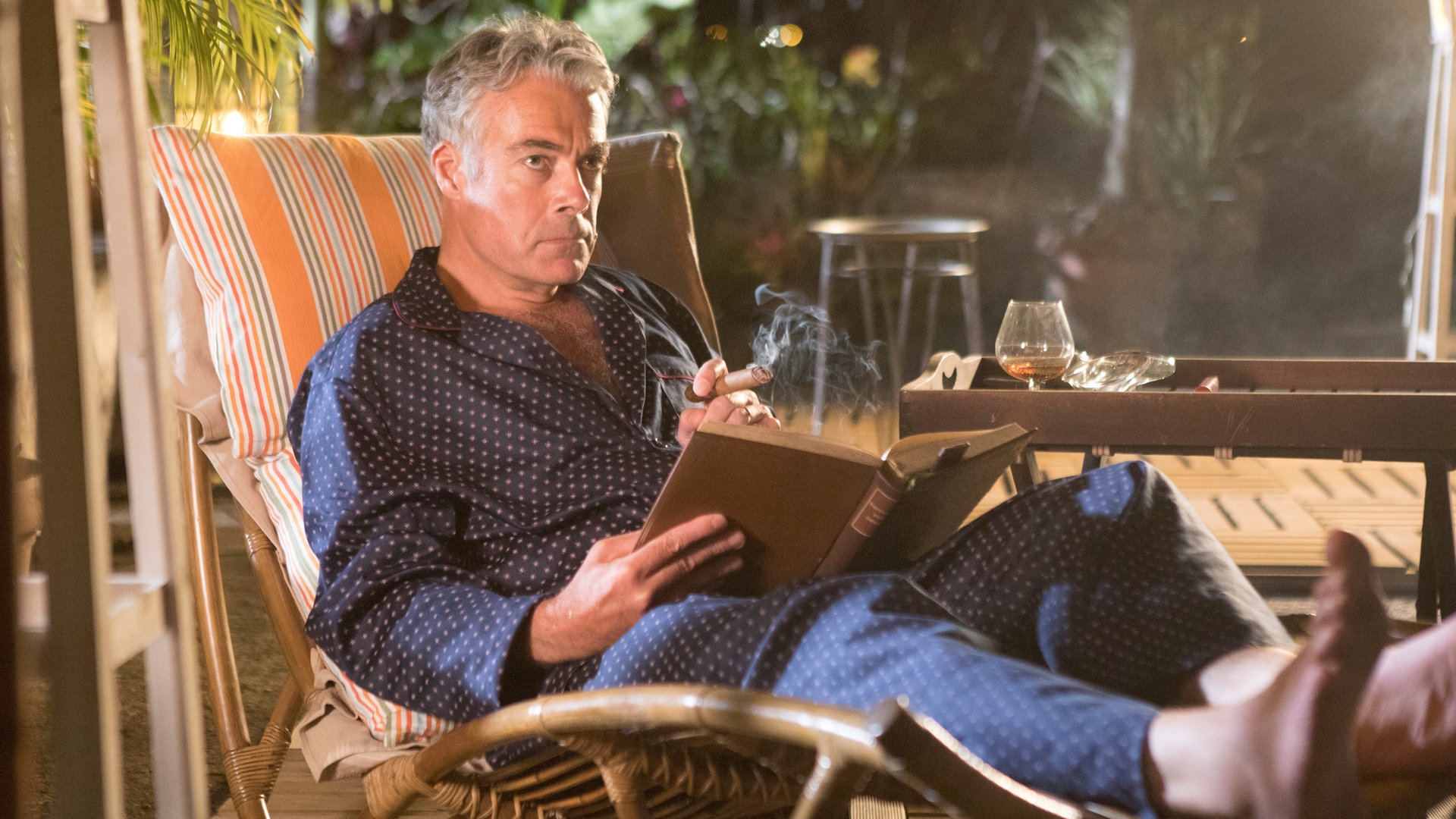 "Death in Paradise - Bittere Bohnen": Am Abend. Benedict Dacre (Robert Portal) sitzt im Bademantel auf einer Bambus-Gartenliege auf seiner Terrasse. Er liest ein Buch und raucht eine Zigarre. Auf dem Tisch steht ein Glas Cognac und ein Aschenbecher.
