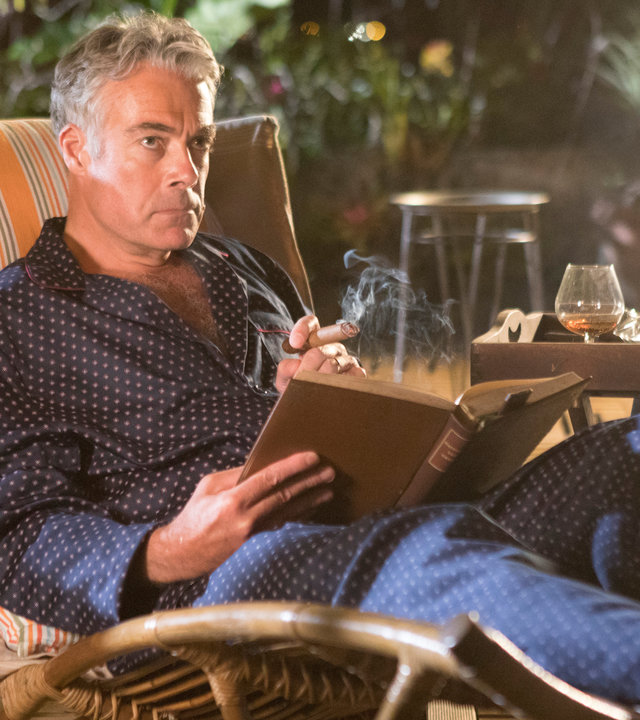 "Death in Paradise - Bittere Bohnen": Am Abend. Benedict Dacre (Robert Portal) sitzt im Bademantel auf einer Bambus-Gartenliege auf seiner Terrasse. Er liest ein Buch und raucht eine Zigarre. Auf dem Tisch steht ein Glas Cognac und ein Aschenbecher.