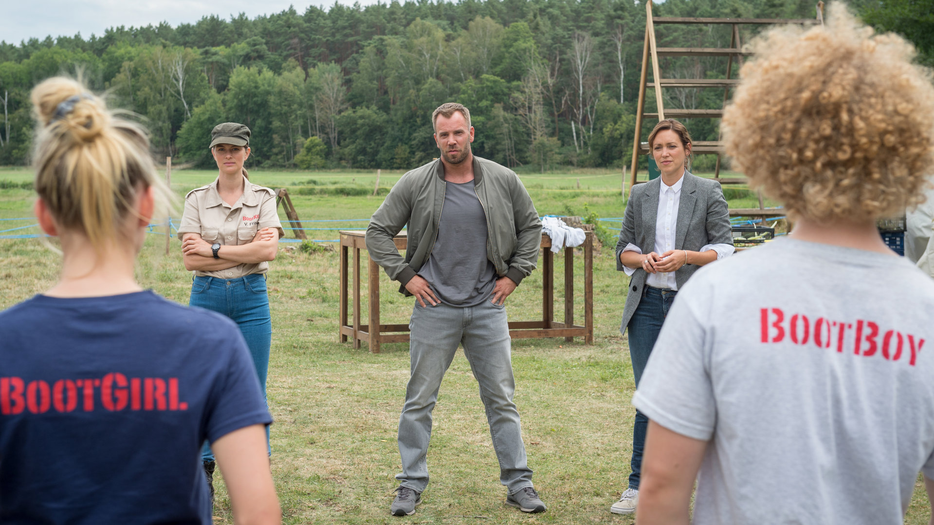 "SOKO Wismar – Zucht und Ordnung": Im Hintergrund sind Geräte für den Hindernislauf des Bootcamps zu sehen. Davor stehen Verhaltenstherapeutin Veronika Frisch (Alexandra Gottschlich), Lars Pöhlmann (Dominic Boeer) und Karoline Joost (Nike Fuhrmann). Diese blicken auf zwei Jugendliche im Bootcamp-Outfit, die mit dem Rücken zur Kamera stehen.