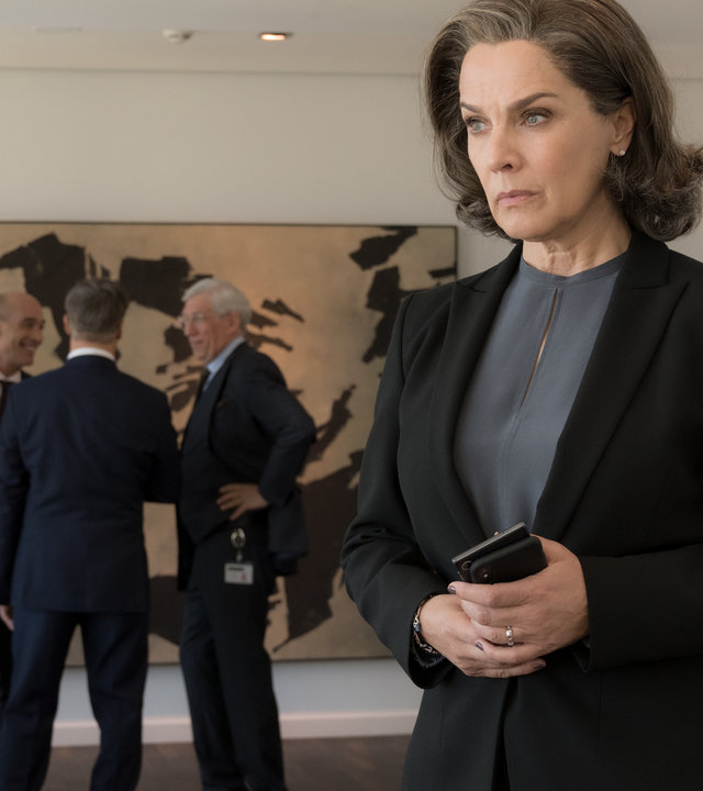 "Bad Banks - Schöne neue Welt": Christelle Leblanc (Désirée Nosbusch) steht in einem großräumigen, hellen Warteraum und schaut nachdenklich vor sich hin. Im Hintergrund diskutiert eine Gruppe von Männern in Anzügen miteinander. Sie machen einen entspannten und gelassenen Eindruck.