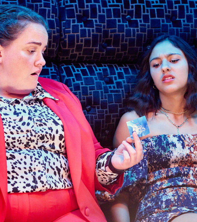 "The Outlaws - Der Dean": Rani (Rhianne Barreto) und Diane (Jessica Gunning) sitzen nebeneinander auf einem Sofa, während Diane Rani ein Kondom hinhält. Beide sind festlich gekleidet.