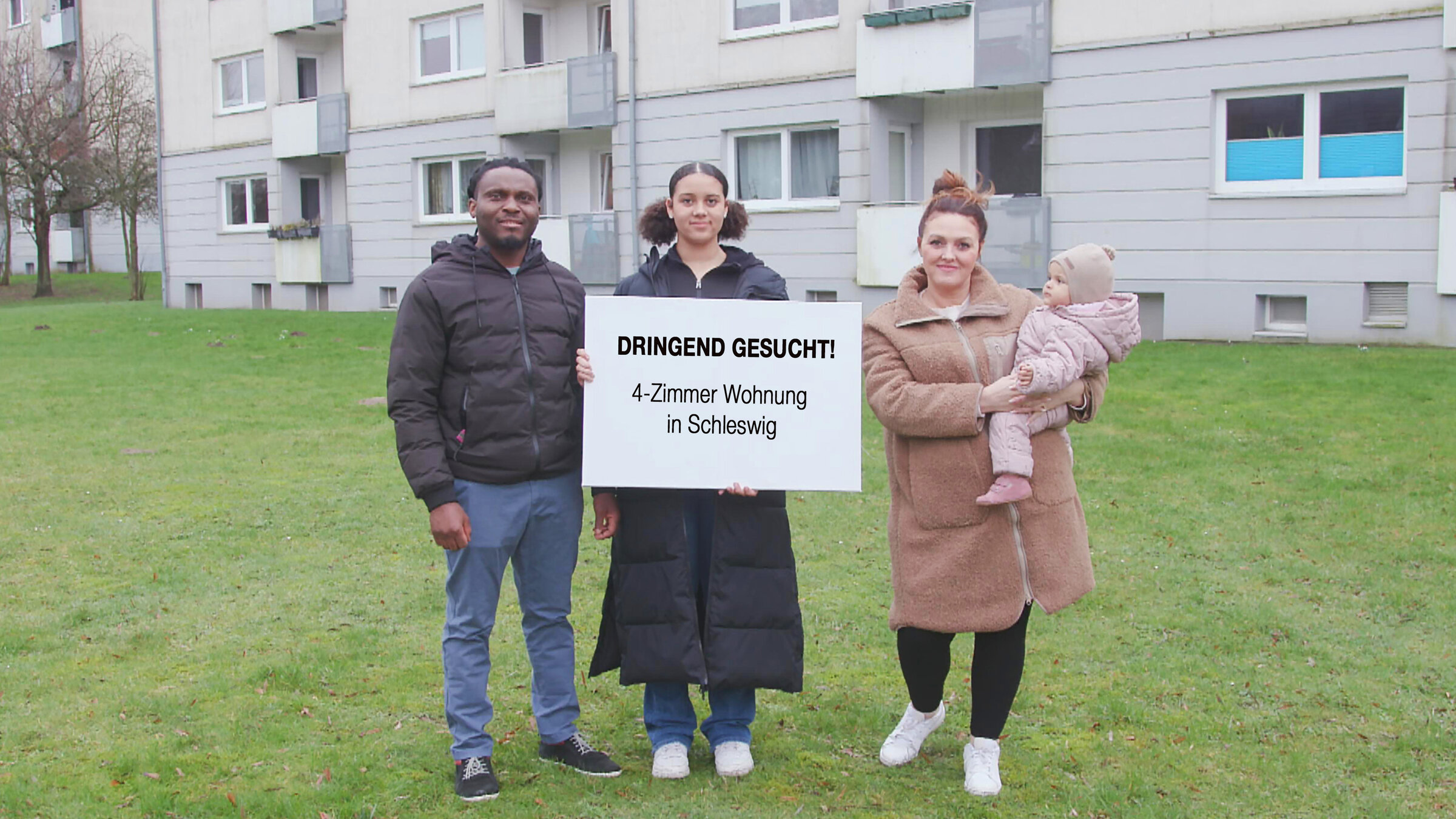 "ZDF.reportage: Albtraum Mieten - Wohnung verzweifelt gesucht": Steven-Ken und Tatyana stehen mit ihren Kindern auf einer Wiese vor einem Wohnblock. Die große Tochter hält ein großes Schild in die Kamera: "Dringend gesucht! 4-Zimmer-Wohnung in Schleswig".
