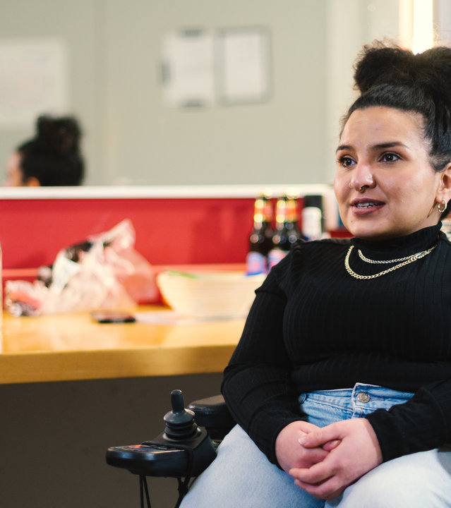 "einfach Mensch - Kübra Sekin: Freie Fahrt für gleiche Rechte": Kübra Sekin, die die Glasknochenkrankheit hat, sitzt in ihrem Rollstuhl vor einem Tisch mit Spiegel und schaut nach links aus dem Bild.