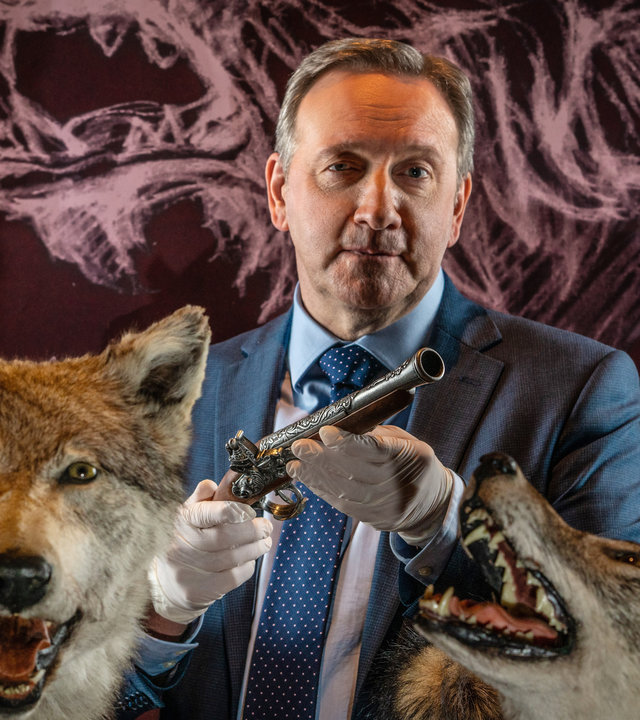 "Inspector Barnaby - Das Wolfmonster von Little Worthy": DCI John Barnaby (Neil Dudgeon) steht zwischen zwei präparierten Wölfen. Über seine Hände hat er Latex-Handschuhe gezogen und hält mit ihnen eine antike Vorderlader-Pistole vor sich.