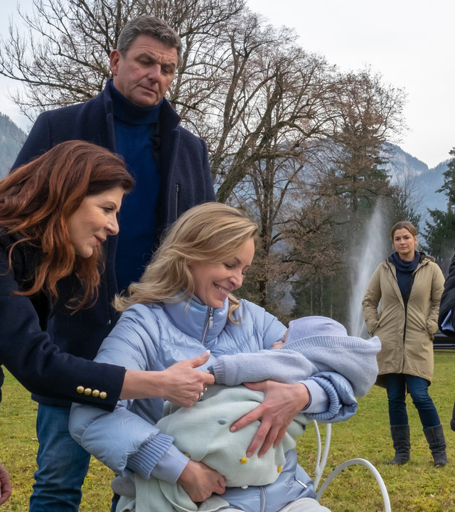 "Der Bergdoktor: Im Frieden (2)": Im Garten auf einem Stuhl sitzend hält Franziska (Simon Hanselmann) ihren Sohn lächelnd im Arm. Thea (Claudia Wenzel) beugt sich über beide und hält die Hand ihres Enkels. Um die beiden herum stehen Martin (Hans Sigl) und Dr. Herold (Lutz Blochberg). Anne (Ines Lutz) steht, traurig blickend, im Hintergrund und beobachte die Szene.