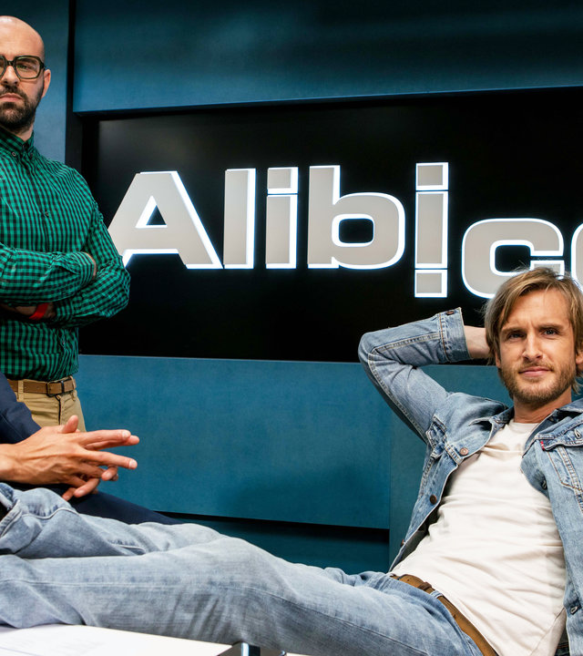 "Alibi.com": Grégory (Philippe Lacheau) sitzt, mit den Füßen auf dem Schreibtisch, vor seinem riesigen Firmenlogo, im Büro. Seine Mitarbeiter Augustin (Julien Arruti) und Mehdi (Tarek Boudali) stehen neben ihm.