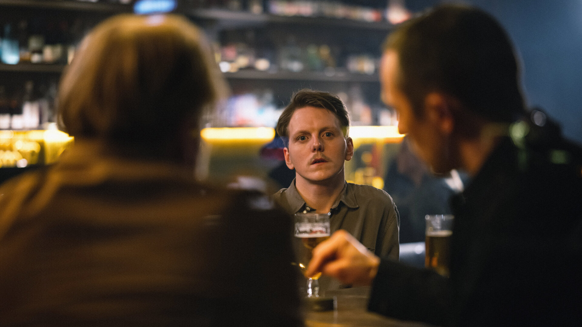 "Toxic Tom - Audun und das Koma der Geilheit": Der schmächtige Audun (Jonas Strand Gravli, M.) mit kurzen braunen Haaren und einem braunen Oberlippenbart blickt abwesend nach vorne, wo ihm zwei Männer (Komparsen) an einem Kneipentisch gegenübersitzen.