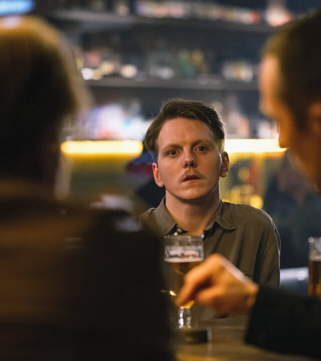 "Toxic Tom - Audun und das Koma der Geilheit": Der schmächtige Audun (Jonas Strand Gravli, M.) mit kurzen braunen Haaren und einem braunen Oberlippenbart blickt abwesend nach vorne, wo ihm zwei Männer (Komparsen) an einem Kneipentisch gegenübersitzen.