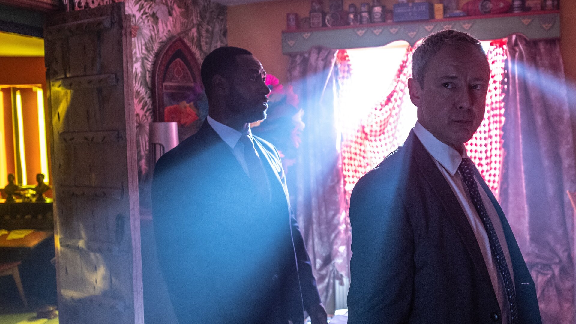 "Detective Grace - Dich will ich töten": DS Glenn Branson (Richie Campbell) und DS Roy Grace (John Simm) stehen nebeneinander in einem unaufgeräumten Zimmer und schauen sich um. Durch das mit rot-weiß-karierten Tüchern abgehängte Fenster scheinen Lichtstreifen von außen herein, die die Szene gespenstisch beleuchten.