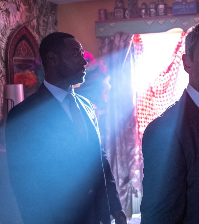 "Detective Grace - Dich will ich töten": DS Glenn Branson (Richie Campbell) und DS Roy Grace (John Simm) stehen nebeneinander in einem unaufgeräumten Zimmer und schauen sich um. Durch das mit rot-weiß-karierten Tüchern abgehängte Fenster scheinen Lichtstreifen von außen herein, die die Szene gespenstisch beleuchten.