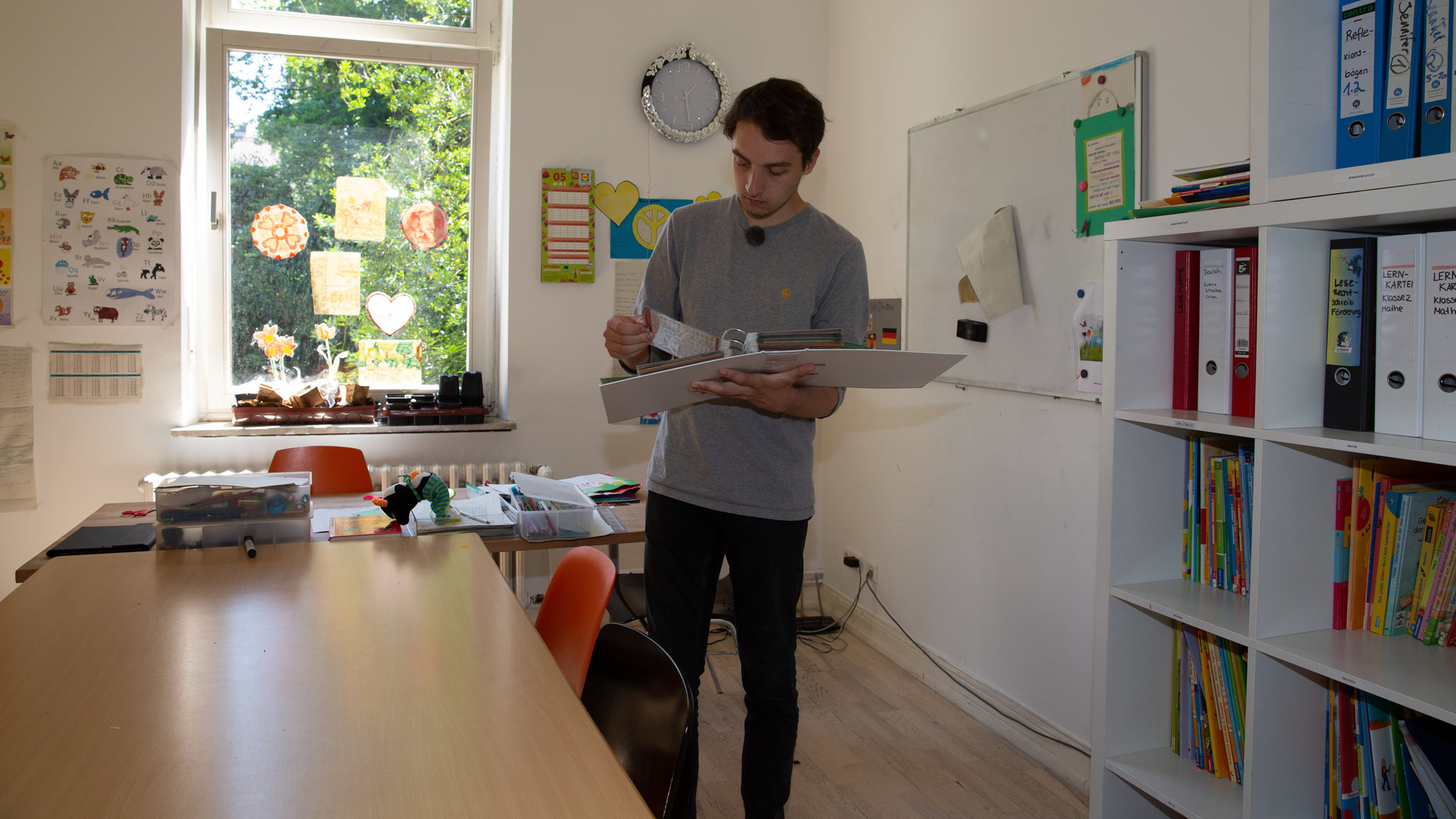 "einfach Mensch - Till Hölzl: Ich tausche Bildung für Wohnung": Till Hölzl in einem Zimmer mit Buchregalen und einem Holztisch auf der linken Seite.