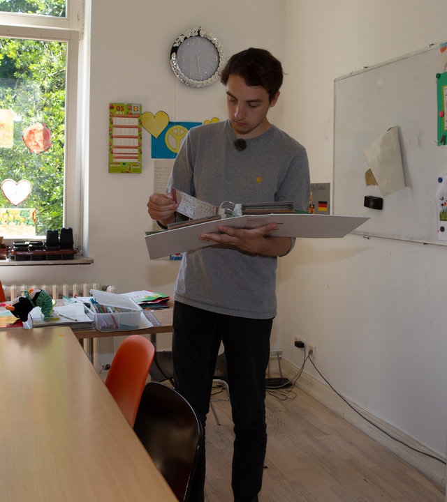 "einfach Mensch - Till Hölzl: Ich tausche Bildung für Wohnung": Till Hölzl in einem Zimmer mit Buchregalen und einem Holztisch auf der linken Seite.