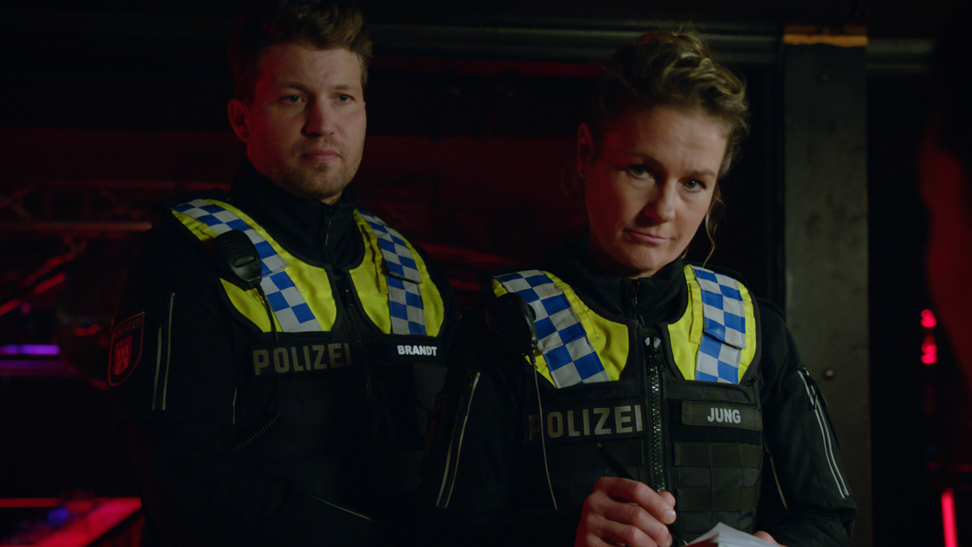 "Notruf Hafenkante - Letzter Einsatz": Der Polizist Nick Brandt (Raúl Richter) und seine Kollegin Franzi Jung (Rhea Harder-Vennewald) stehen nebeneinander in einem Club. Franzi hält ein Notizbuch und einen Stift in den Händen und blickt ernst auf etwas vor sich.
