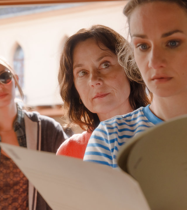 "Mit Herz und Holly - Muttergefühle": In einer Arztpraxis hält Dr. Holly Sass (Karoline Teska) eine Akte in der Hand und liest mit angespanntem Gesichtsausdruck darin. Dr. Katrin Herz (Inka Friedrich) steht dicht neben ihr und beobachtet sie aufmerksam. Im Hintergrund wartet Dana Kral (Ina Hout) mit genervtem Gesichtsausdruck.