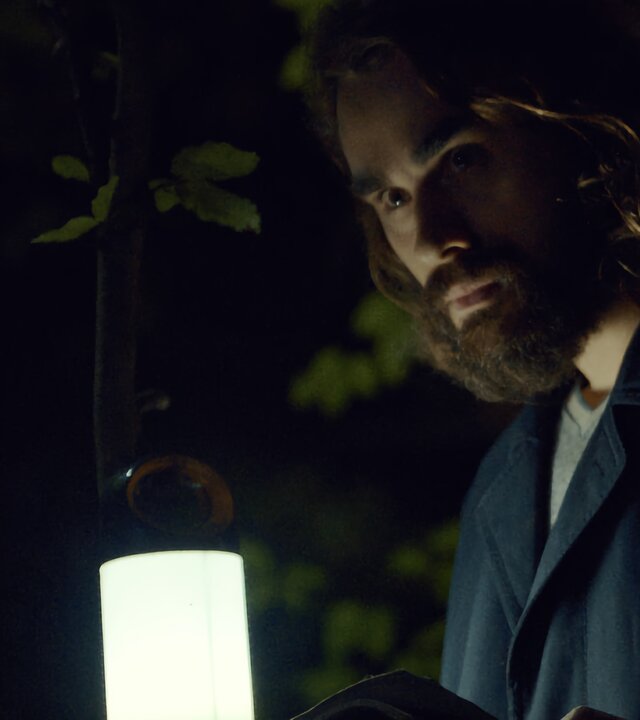 "Moresnet - The Book of Death: Das Tagebuch": Ben Schotz (Boris Van Severen) steht nachts in einem Wald. In seiner linken Hand hält er ein aufgeschlagenes Tagebuch, das er mit einer elektrischen Laterne beleuchtet, die er in seiner rechten Hand hält. Die Szene wirkt durch den fahlen Lichtschein gespenstisch.