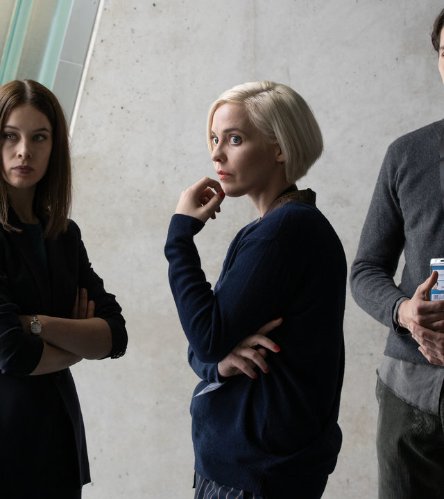 "Bad Banks - Kollateralschaden": Jana (Paula Beer), Ben (Noah Saavedra) und Lilou Guerin (Elisabet Johannesdottir) stehen wartend in einer Ecke vor einer modernen Beton- und Glaswand. Alle schauen angespannt in die gleiche Richtung.