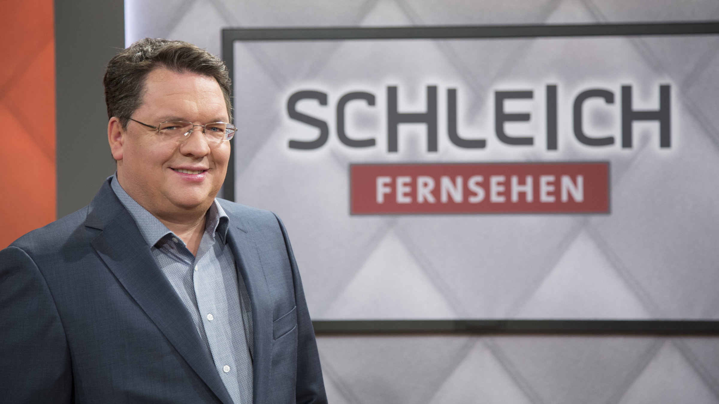 "SchleichFernsehen": Helmut Schleich (nur Oberkörper) vor einem Hintersetzer, auf dem "Schleich Fernsehen" steht.