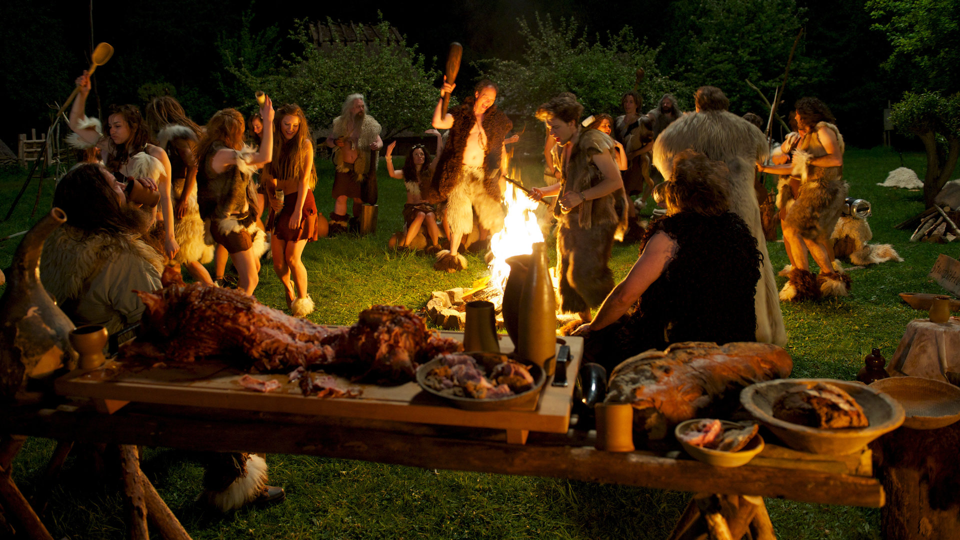 "SOKO Wismar - Neandertal ist überall": Um ein Lagerfeuer herum stehen viele Menschen in Fell und Leder gekleidet. Sie trinken, essen und feiern. Im Vordergrund ist ein Festmahl aus Fleisch und Brot auf einem Holztisch aufgebaut.