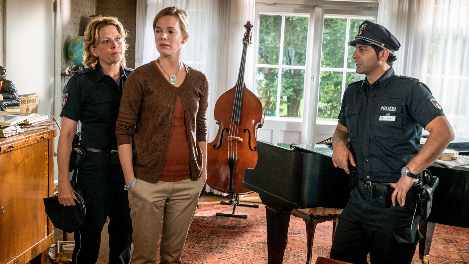 "Notruf Hafenkante - Hausmusik": Die Polizistin Claudia Fischer (Janette Rauch) steht seitlich hinter Constanze Magnus (Milena Dreissig) und sieht sie an. Constanze hat beide Hände in den Hosentaschen und schaut nachdenklich zur Seite. Neben den beiden Frauen steht Claudias Kollege Tarik Coban (Serhat Cokgezen). Er lehnt auf einem schwarzen Flügel. Im Hintergrund steht ein Kontrabass.