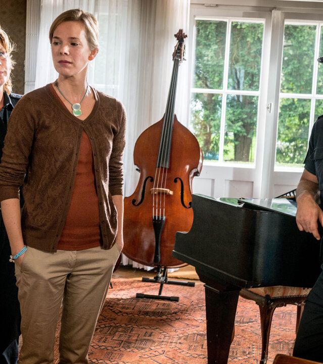 "Notruf Hafenkante - Hausmusik": Die Polizistin Claudia Fischer (Janette Rauch) steht seitlich hinter Constanze Magnus (Milena Dreissig) und sieht sie an. Constanze hat beide Hände in den Hosentaschen und schaut nachdenklich zur Seite. Neben den beiden Frauen steht Claudias Kollege Tarik Coban (Serhat Cokgezen). Er lehnt auf einem schwarzen Flügel. Im Hintergrund steht ein Kontrabass.