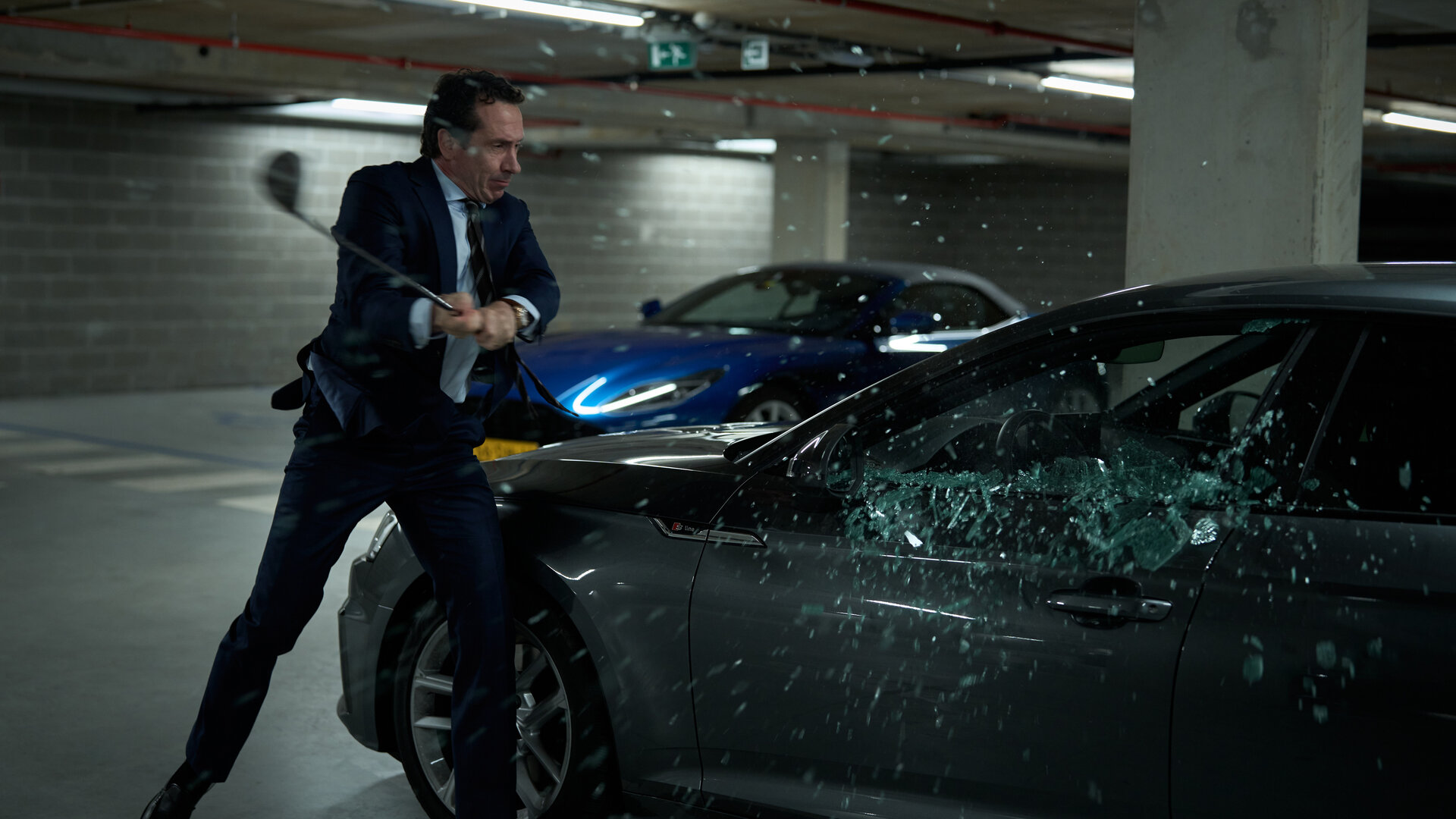 "Elixir – The Pain Killers - Das Versprechen": In einem Parkhaus schlägt Juliën (Jacob Derwig) mit einem Golfschläger auf ein dort abgestelltes Auto ein. Die Scheibe des Fensters auf der Fahrerseite zerspilttert gerade.