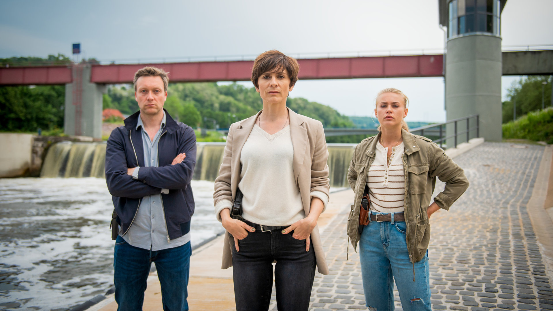 "In Wahrheit – Unter Wasser": Die Ermittler Freddy Breyer (Robin Sondermann), Judith Mohn (Christina Hecke), Lisa (Jeanne Goursaud) alle mit Kamerablick am Flussufer der Nied.