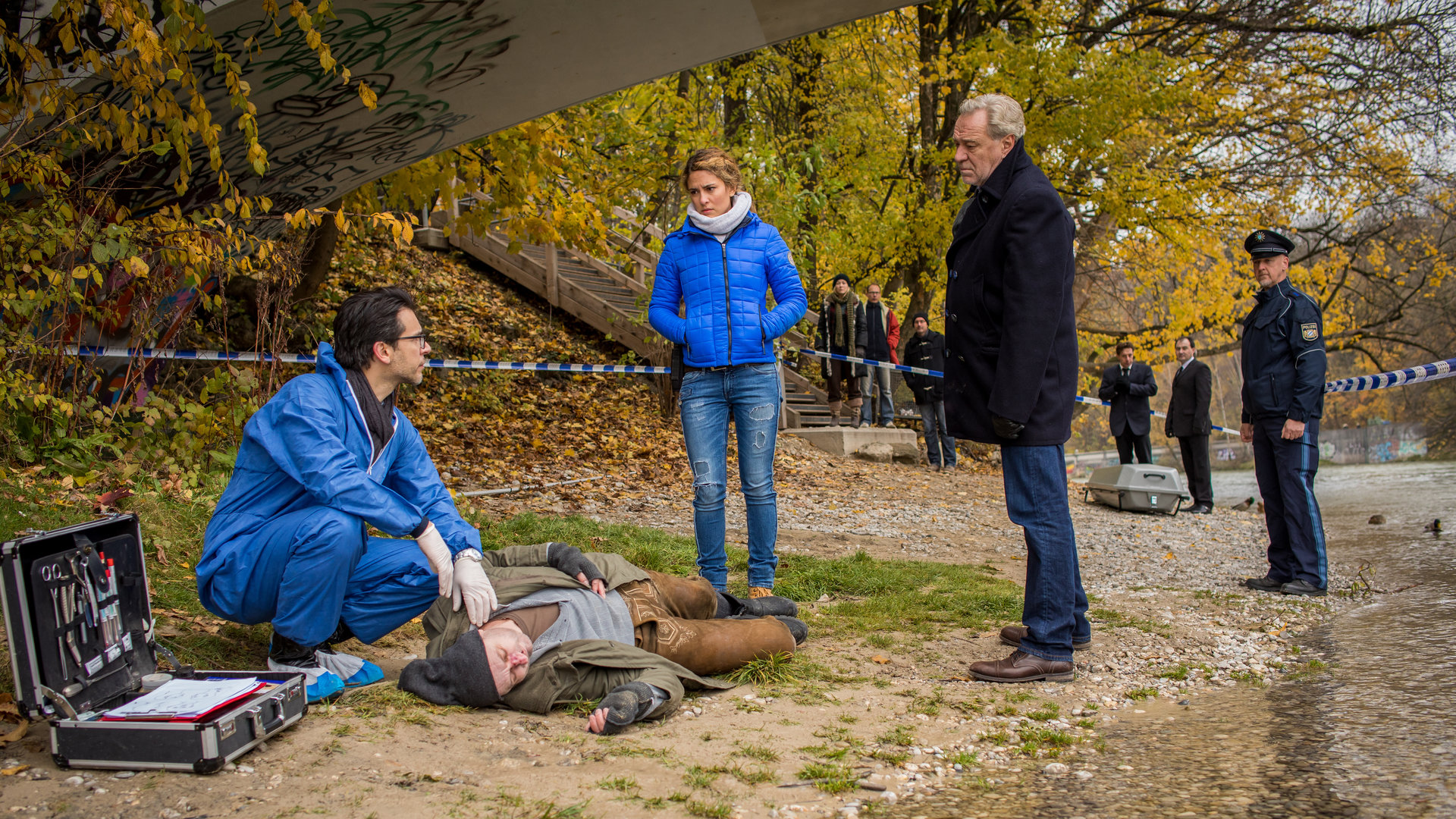 "SOKO München - Tod unter Brücken": Am Ufer unter einer Brücke liegt eine Leiche. Dr. Weissenböck (Florian Odendahl) sitzt in der Hocke daneben, er hat seine linke Hand am Hals der Leiche und schaut zu Arthur Bauer (Gerd Silberbauer) und Toni Bischoff (Amanda da Gloria). Im Hintergrund stehen Schaulustige (Komparsen), einen Polizisten und zwei Bestatter mit einem Sarg.