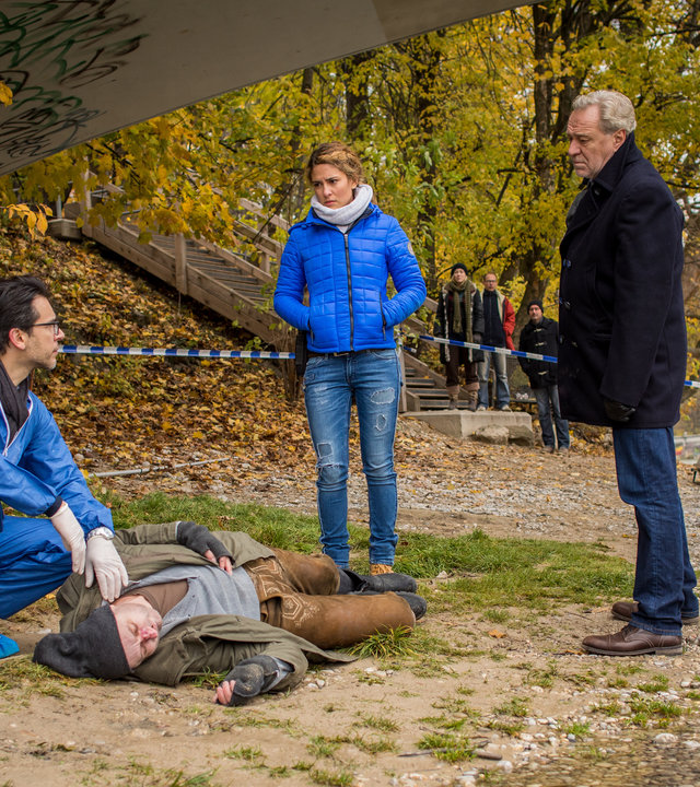 "SOKO München - Tod unter Brücken": Am Ufer unter einer Brücke liegt eine Leiche. Dr. Weissenböck (Florian Odendahl) sitzt in der Hocke daneben, er hat seine linke Hand am Hals der Leiche und schaut zu Arthur Bauer (Gerd Silberbauer) und Toni Bischoff (Amanda da Gloria). Im Hintergrund stehen Schaulustige (Komparsen), einen Polizisten und zwei Bestatter mit einem Sarg.