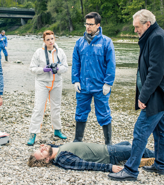 "SOKO München - Von Luft und Liebe": Das Opfer liegt tot auf den Steinen eines trockenen Flussbetts. Arthur Bauer (Gerd Silberbauer) steht neben ihm und blickt zu Resi Schwaiger (Mersiha Husagic), die gerade telefoniert. Zwischen den beiden stehen Dr. Weissenböck (Florian Odendahl) und Billie Curio (Sina Reiß). Im Hintergrund stochern zwei Spurensicherer (Komparsen) im Fluss.