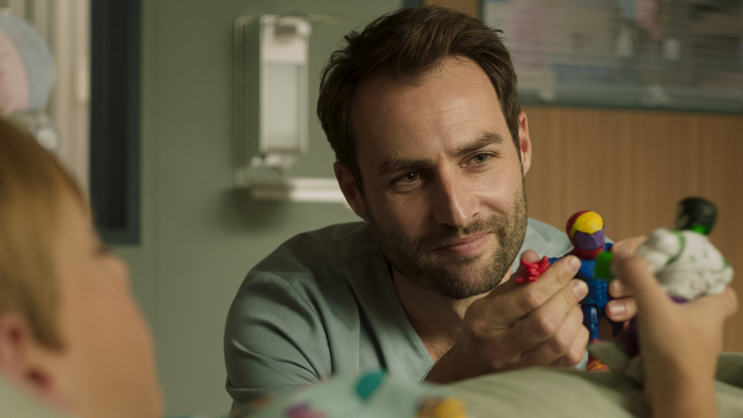 "Doktor Ballouz - Blutsbrüder": Dr. Schilling (Daniel Fritz) sitzt am Krankenbett seines Patienten Leon (Magnus Alexander Jónsson) und spielt mit ihm mit seinen Actionfiguren.