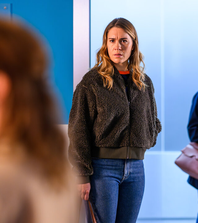"Bettys Diagnose - Wanderjahre": Betty (Henrike Hahn) steht vor der offenen Aufzugstür und schaut verdutzt zu Prof. Dr. Ariane Schönfelder (Dagny Dewarth) hinüber. Zoe Breitner (Catherine Chikosi) steht neben Betty und schaut ebenfalls zu Ariane Schönfelder hinüber.
