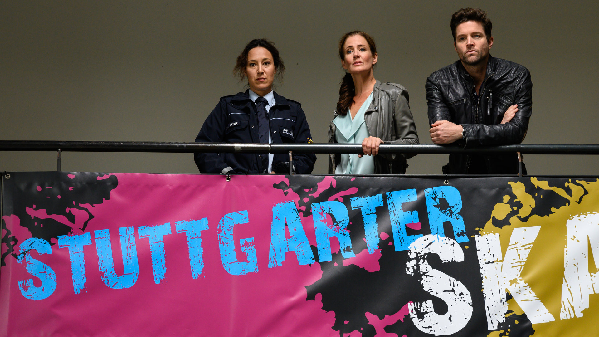 "SOKO Stuttgart - skate or die": Sibylle Beyer (Bärbel Stolz), Selma Kirsch (Yve Burbach) und Jo Stoll (Peter Ketnath) stehen nebeneinander am Geländer der Empore in der Skatehalle und schauen in die Halle hinunter. Unter ihnen hängt ein großes Banner, das für den Skate-Contest wirbt.
