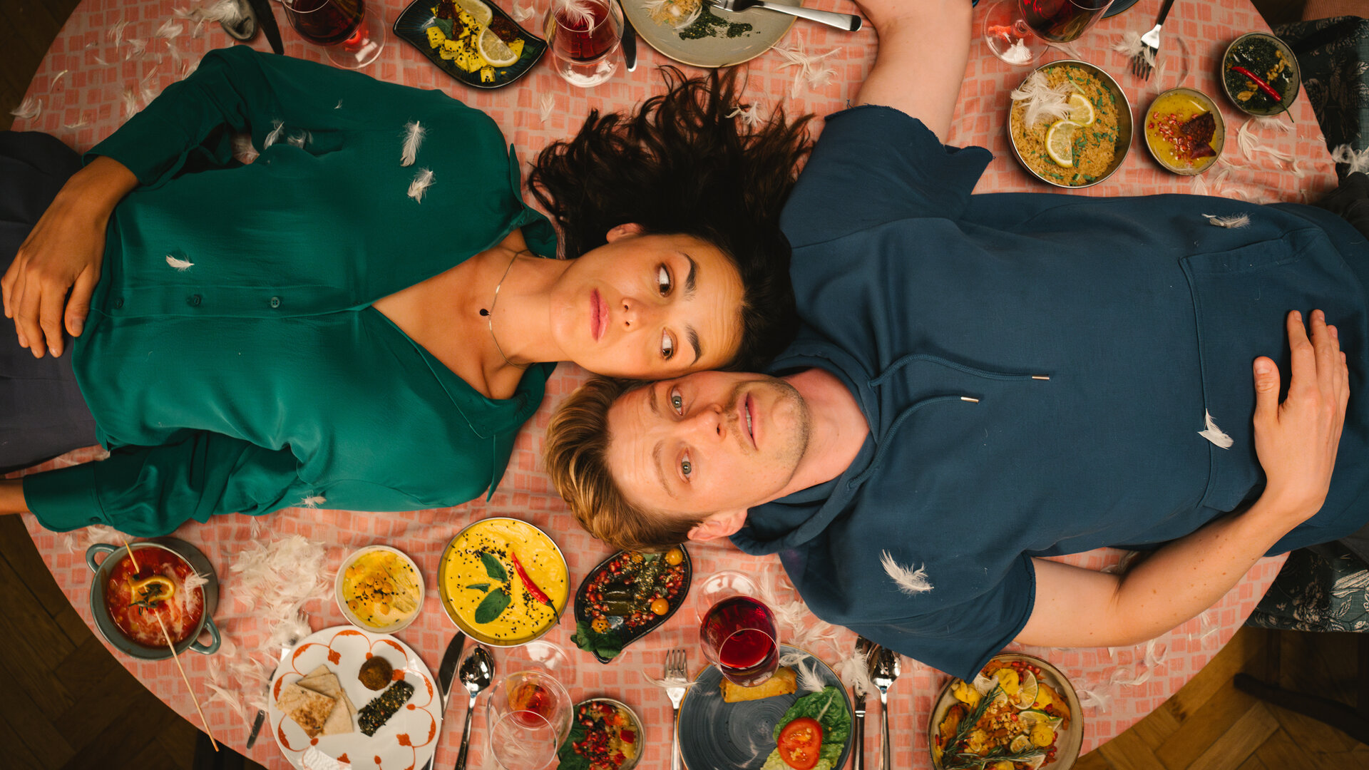 "Feste Feiern": Blick von oben: Knut (Helgi Schmid) und Karo (Lucie Heinze) liegen auf dem Rücken und mit den Köpfen nebeneinander auf einem gedeckten Tisch.
