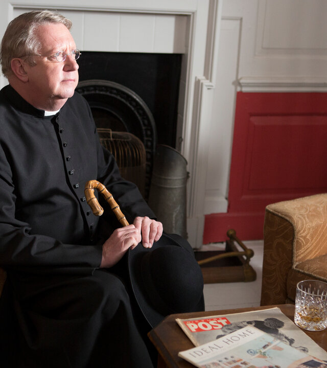 "Father Brown - Die Leichenräuber": Father Brown (Mark Williams) sitzt nachdenklich am Kamin auf einem Sessel.