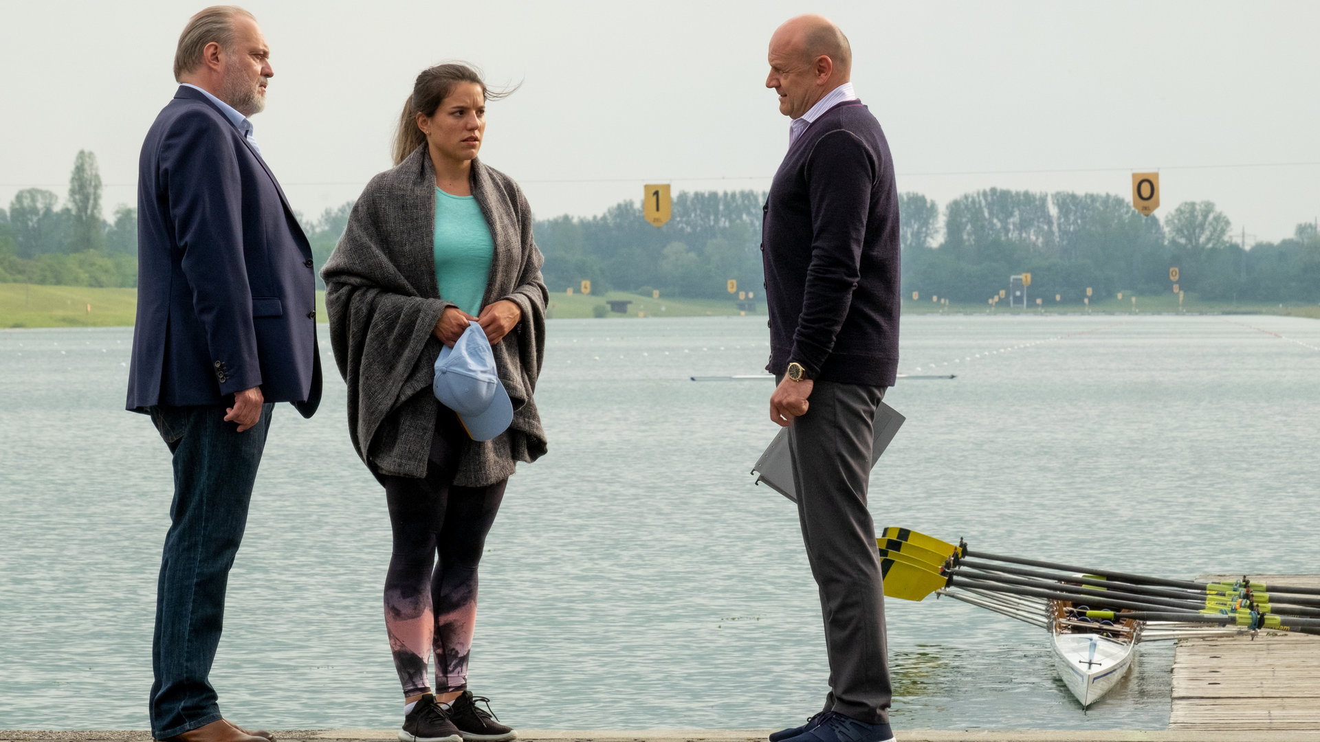 "Der Alte - Der schwarze Tunnel": Hauptkommissar Richard Voss (Jan-Gregor Kremp) steht vor der Ruderregatta-Strecke und spricht mit Julia Eich (Mona Vojacek Koper), die eine Decke um ihre Schultern gelegt hat und Werner Thieme (Oliver Stokowski). Im Hintergrund sieht man einen Bootssteg, an dem ein Sport-Ruderboot festgemacht ist.