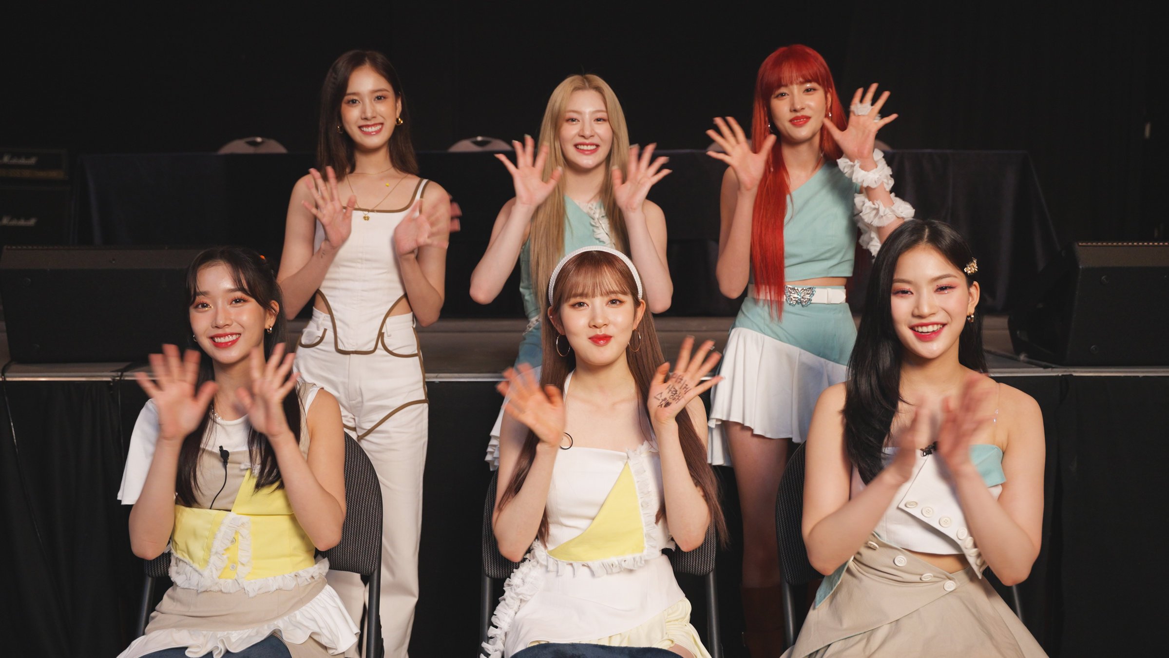 "Kultur-King Korea - Wie südkoreanische Popkultur die Welt erobert": Sechs asiatische, junge Frauen. Drei vorne sitzend, drei dahinter stehend. Sie lächeln in die Kamera und winken mit beiden Händen. Sie haben alle lange glatte Haare, drei davon schwarze, eine braune, eine blonde und eine rote. Sie tragen roten Lippenstift. Die Kleidung ist vornehmlich weiß mit teilweise gelben, türkisen und hellbraunen Akzenten.