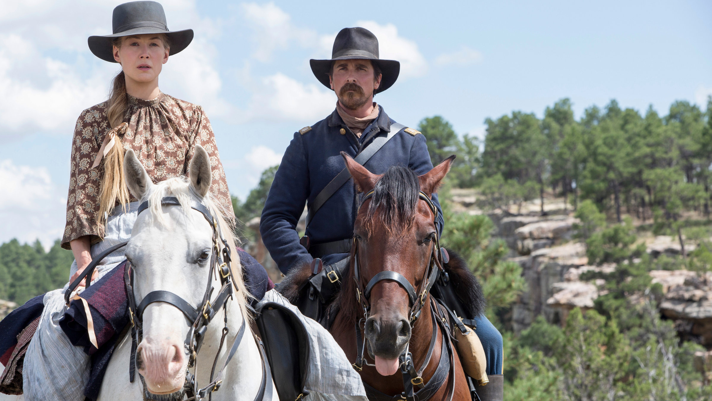 "Feinde - Hostiles": Rosalee (Rosamund Pike) und Captain Blocker (Christian Bale) sitzen auf ihren Pferden und reiten hintereinander durch offenes Gelände.