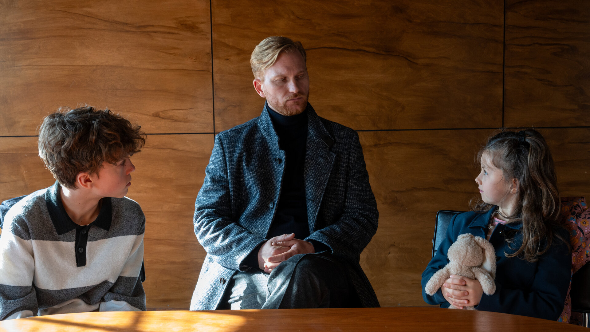 "SOKO Stuttgart - Rosenkrieg": Sven Stengle (Lucas Prisor) sitzt zwischen seinen beiden Kindern Charles (Finn Hubel) und Louise (Sophia Pfeifer) im Befragungsraum des Präsidiums. Er schaut streng auf seine Tochter, die mit ihren Händen fest einen Kuscheltierhasen umklammert.