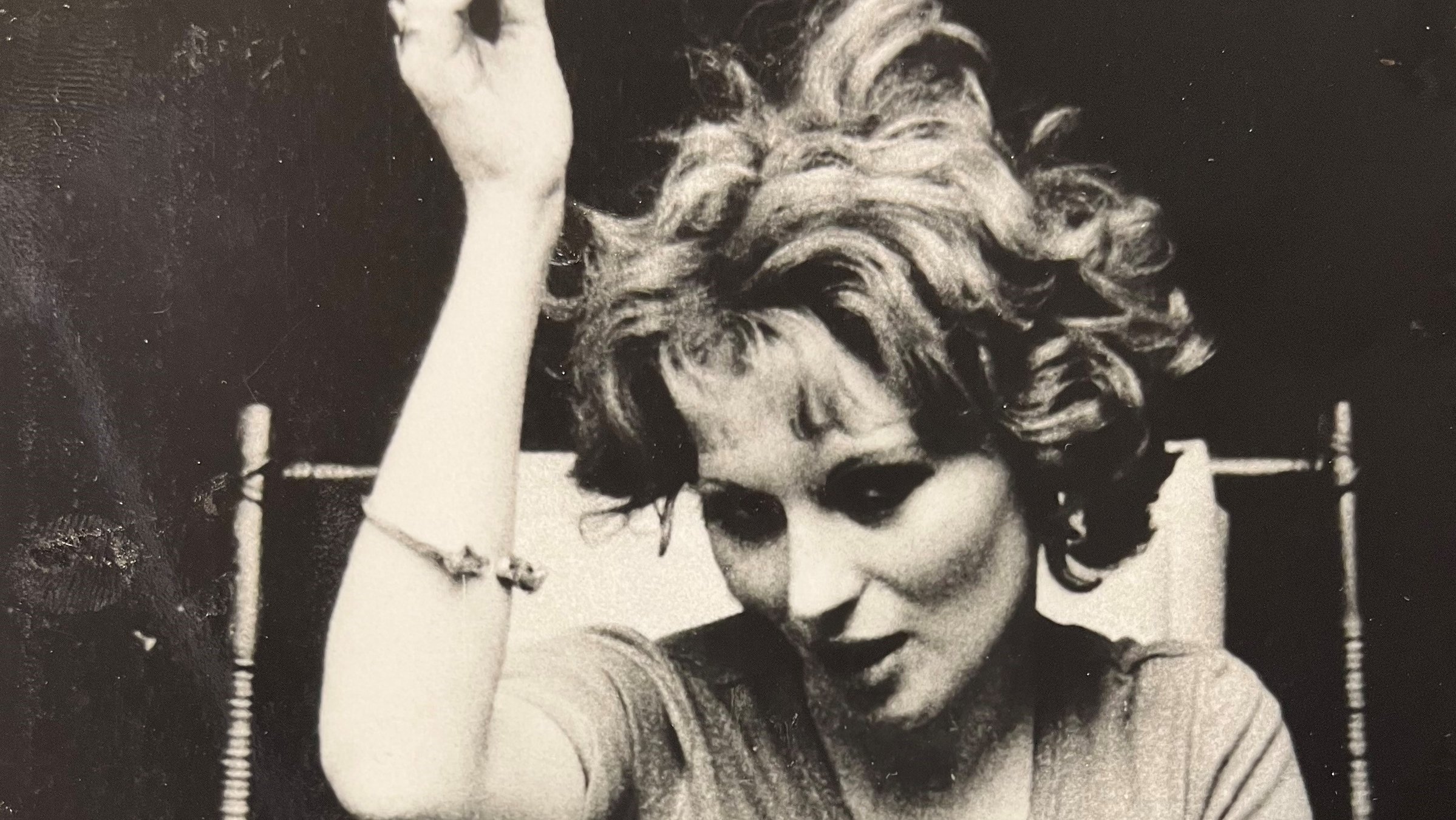 "Ein ganz und gar verwahrlostes Mädchen": Schwarz-Weiß-Aufnahme: Rita (Rita Rischak) sitzt auf einem Stuhl und hebt den rechten Arm nach oben. Der linke liegt auf ihrem Bein. Sie hat den Kopf zur Seite geneigt und sieht in die Kamera.