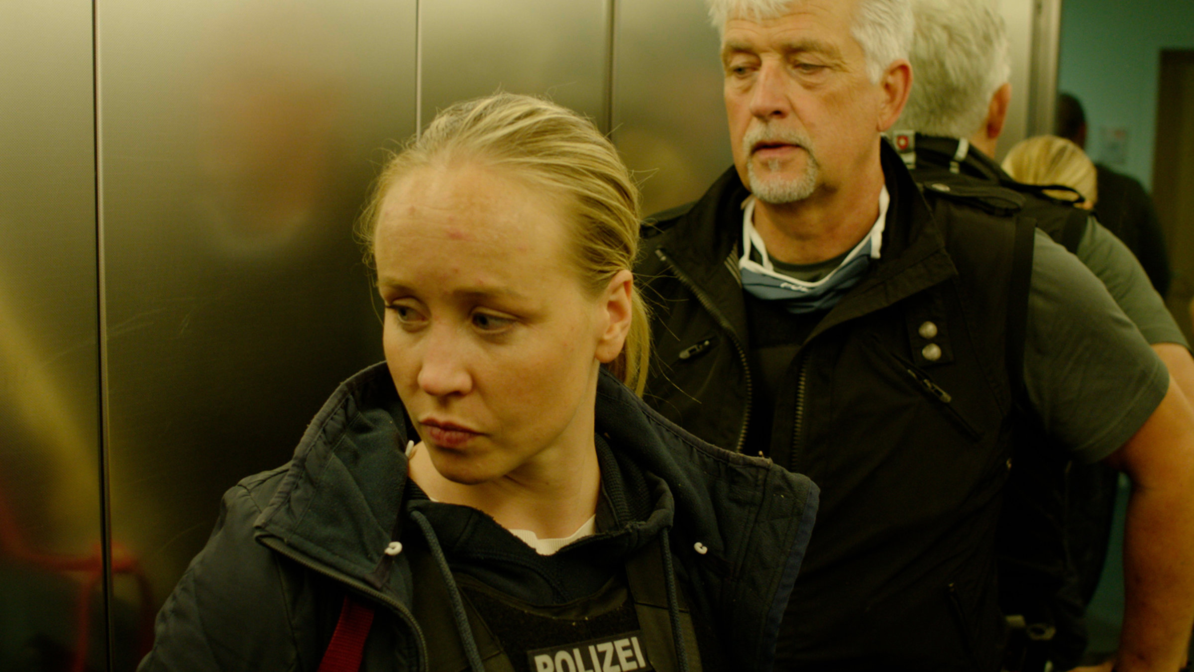 "37°: Auf der Spur der Täter - Delikt Kinderpornografie": Eine junge blonde Frau und ein weißhaariger Mann in Polizeiweste stehen im Fahrstuhl vor der Tür.