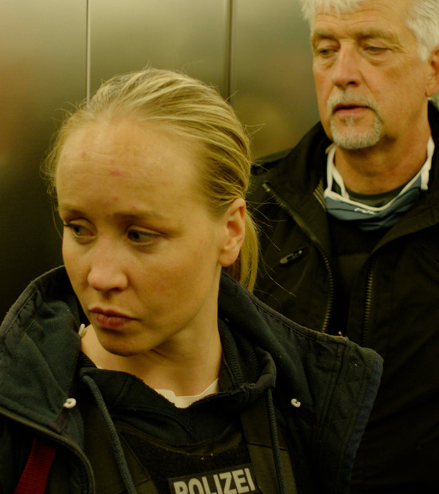"37°: Auf der Spur der Täter - Delikt Kinderpornografie": Eine junge blonde Frau und ein weißhaariger Mann in Polizeiweste stehen im Fahrstuhl vor der Tür.