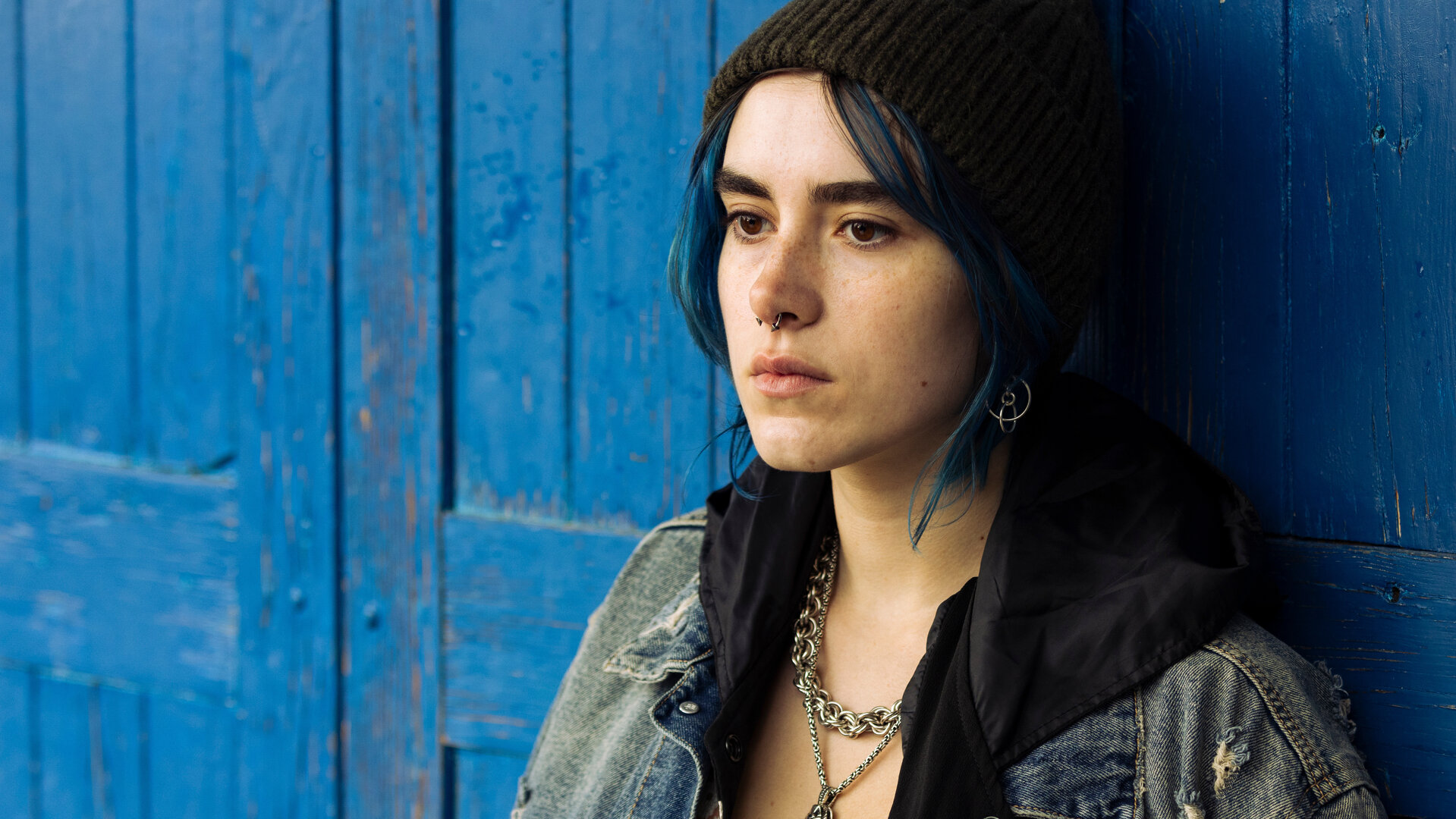 "Phoenix - Change Or Die: Bereit zum Mord": Nina (Alva Schäfer), eine junge Frau mit kinnlangen blau gefärbten Haaren und auffälligem Silberschmeck inklusive Nasenring, steht vor einer blau lackierten Holztüre.