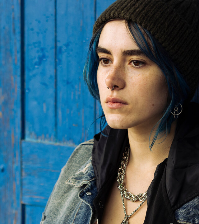 "Phoenix - Change Or Die: Bereit zum Mord": Nina (Alva Schäfer), eine junge Frau mit kinnlangen blau gefärbten Haaren und auffälligem Silberschmeck inklusive Nasenring, steht vor einer blau lackierten Holztüre.