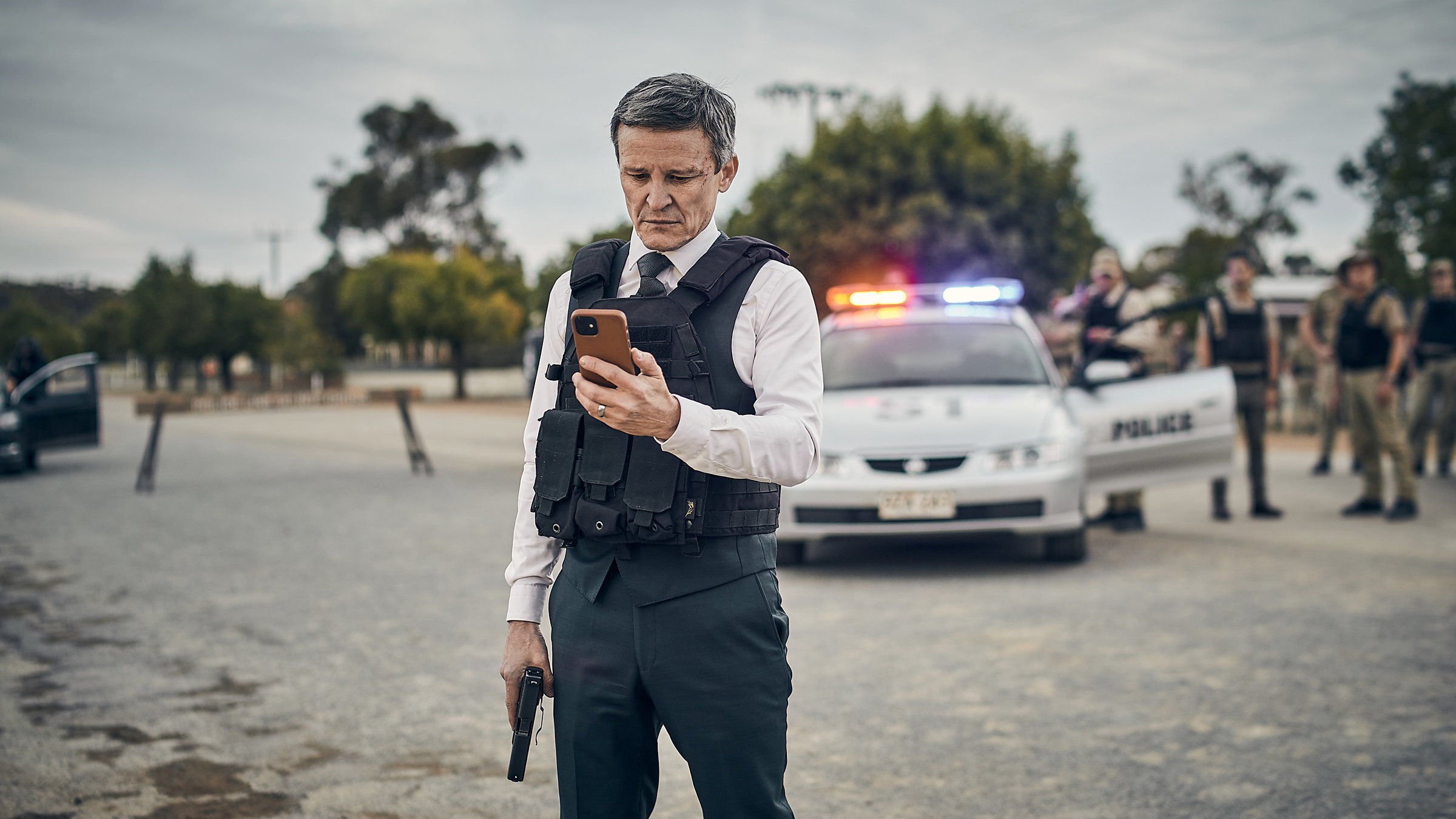 "The Tourist: Burrito, der Erlöser": Detective Inspector Lachlan Rogers (Damon Herriman) steht auf einer abgesperrten Straße. In der einen Hand hält er seine Waffe, in der anderen sein Handy, auf das er schaut. Im Hintergrund sind ein Polizeiwagen und mehrere Polizisten zu sehen.