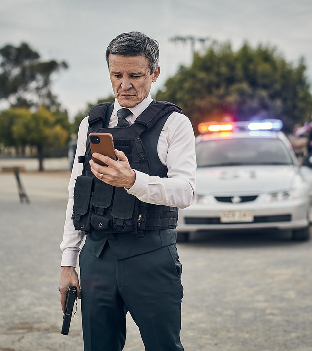"The Tourist: Burrito, der Erlöser": Detective Inspector Lachlan Rogers (Damon Herriman) steht auf einer abgesperrten Straße. In der einen Hand hält er seine Waffe, in der anderen sein Handy, auf das er schaut. Im Hintergrund sind ein Polizeiwagen und mehrere Polizisten zu sehen.