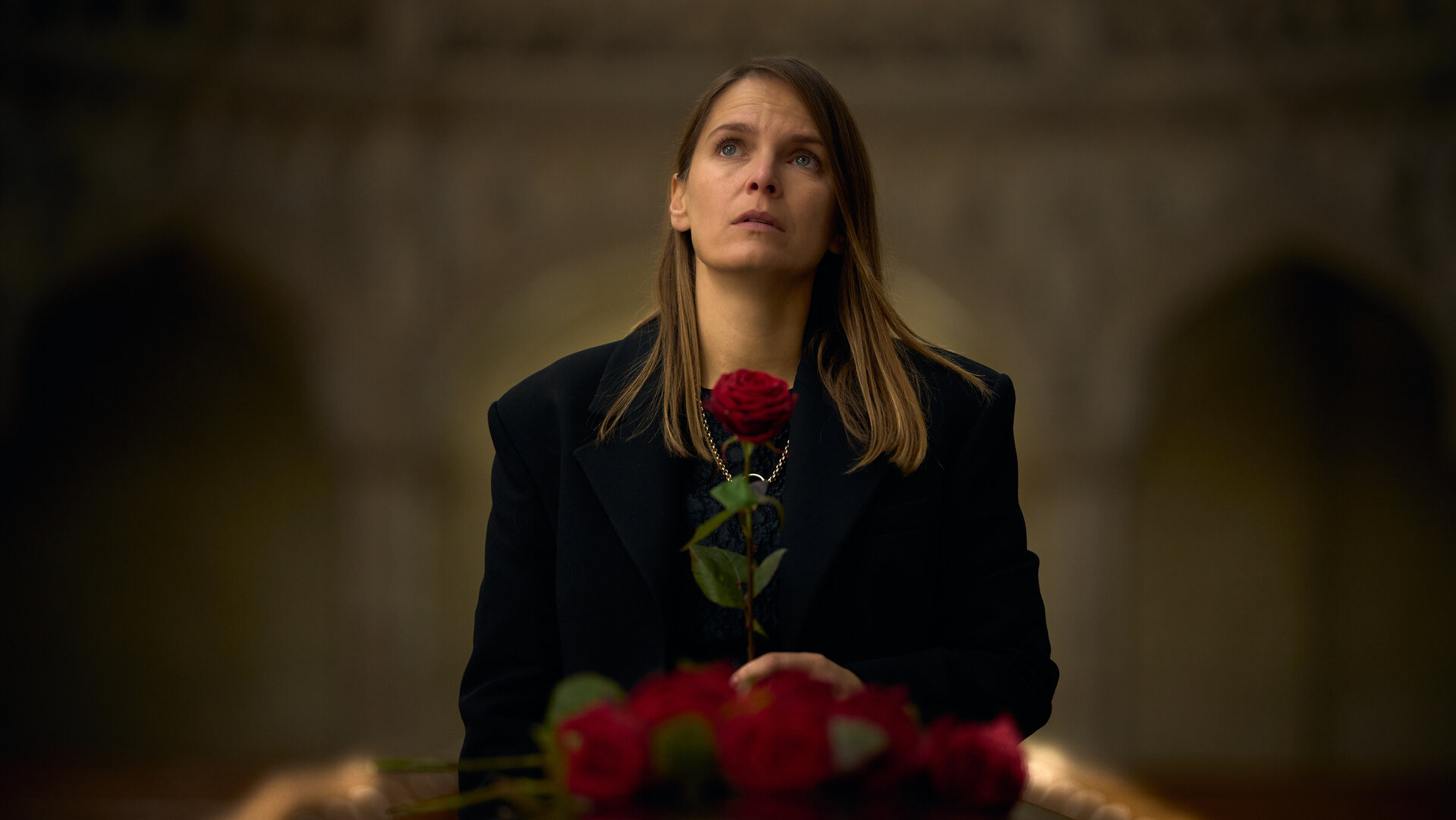 "Elixir – The Pain Killers - Das Gehirn": Isabelle (Hanna Verboom) steht in einer Kirche. Sie ist schwarz gekleidet, schaut nach oben und hält eine rote Rose in der Hand. Vor ihr liegt der Deckel eines Sargs, auf dem weitere Rosen liegen.